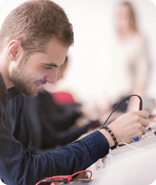 Młody mężczyzna pracuje przy stanowisku elektronicznym, koncentrując się na lutowaniu lub podłączaniu przewodów do urządzenia. W tle rozmyte postacie innych osób, sugerujące pracę zespołową lub szkolenie.