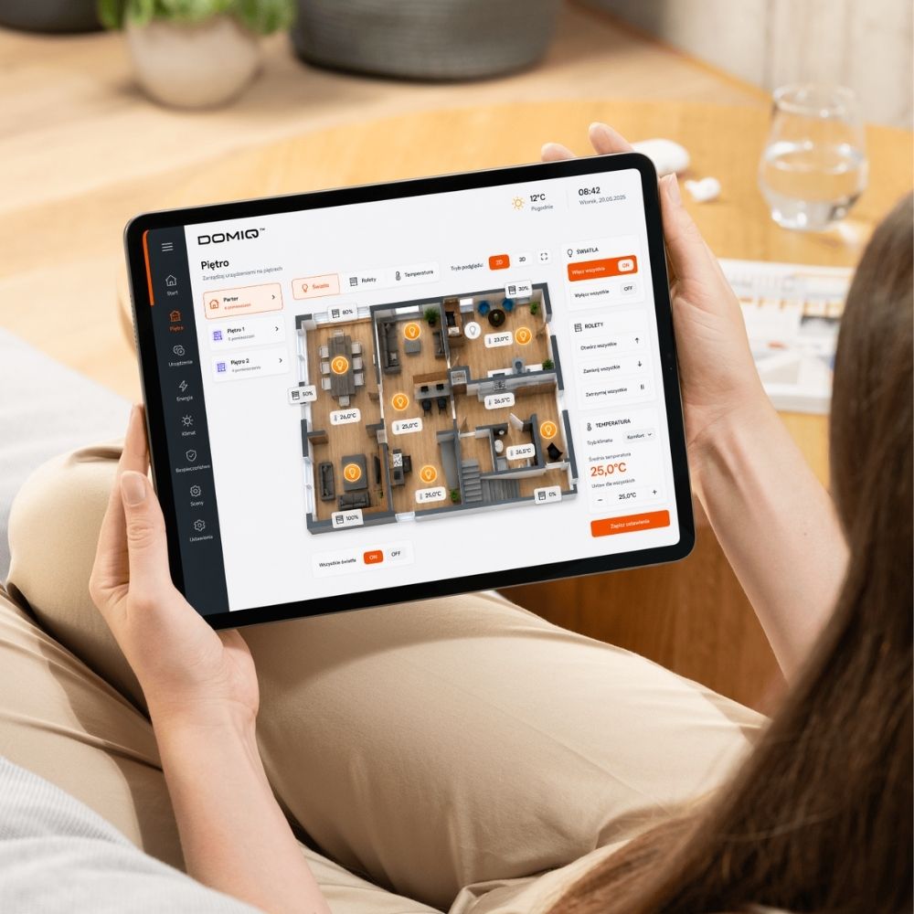 Na zdjęciu osoba która siedząc trzyma na kolanach IPADa któego obserwuje. Na IPADzie wyświetlana jest wizualizacja domu w aplikacji DOMIQ Remote. Ddzięki wizualizacji można szybko sterować systemem smart home.